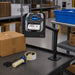 SATO mobile printer mounted in an AgozTech adjustable arm holder at a packing workstation with boxes, tape, and labeling supplies.