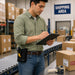Warehouse worker in a sage-green shirt and blue jeans writing on a clipboard in a shipping area while wearing a black AGOZ Datalogic handheld scanner grip holster on his belt.